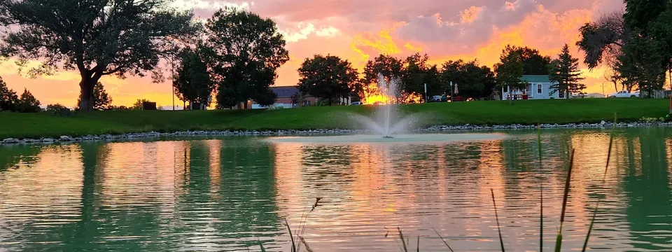 Blue Spruce Pond in Severance at sunset: a fountain on the water, cottonwoods along the far bank and a pink and orange sky